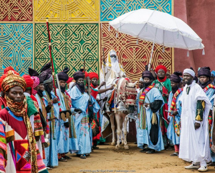 Nigeria, Kano State, Kano. The Emir of Kano mounted on a white horse pauses beside a traditionally decorated wall at his palace with a group of well-dressed followers in attendance.